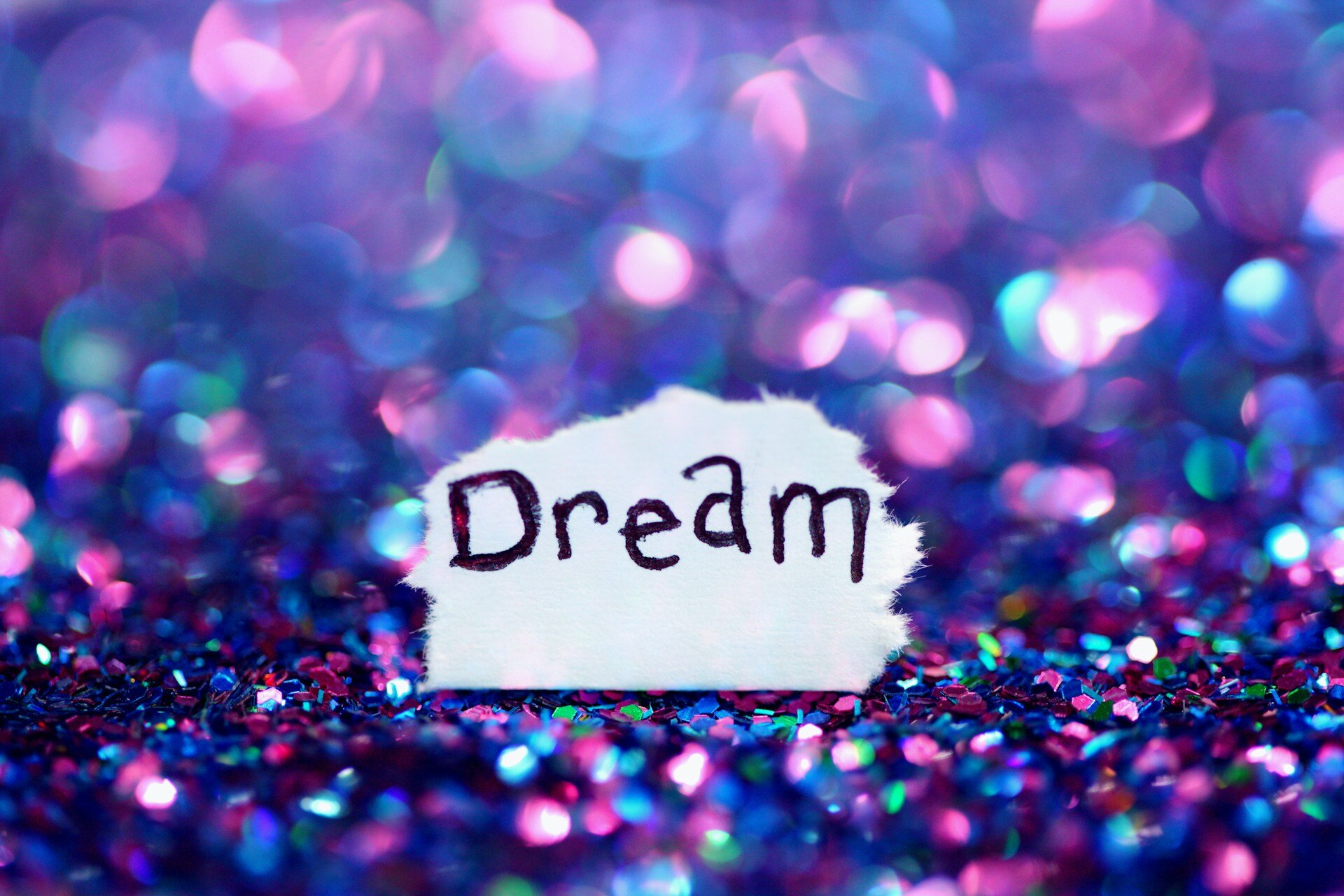 Auf dem Bild sieht man ein Stück Papier, auf dem das Wort "Dream" geschrieben ist. Ringsherum sind blau und violettfarbene Glitzer, die im Hintergrund verschwimmen | © Alexander Grey/Unsplash