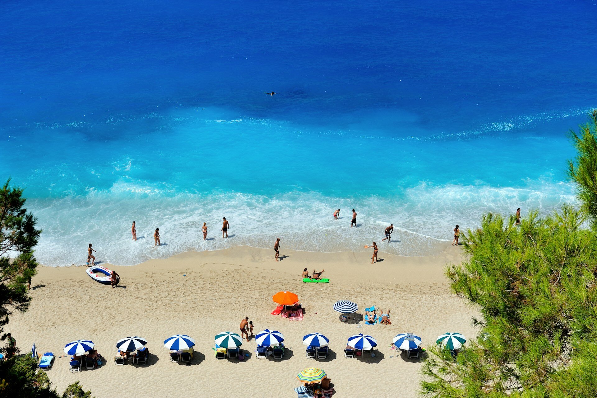 Auf dem Bild sieht man einen Strand mit mehreren blau-weißen und ein paar bunten Sonnenschirmen und Personen, die mit den Füßen im Wasser stehen. Das Meer hat eine tiefblaue Farbe | © Nikos Zacharoulis / Unsplash