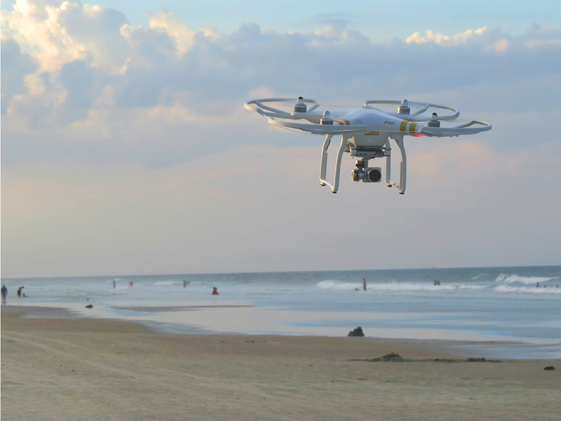 Auf dem Bild sieht man eine Drohne, die am Strand fliegt. Im Hintergrund sieht man das Meer und ein paar Personen | © Matt Pritchard / Unsplash