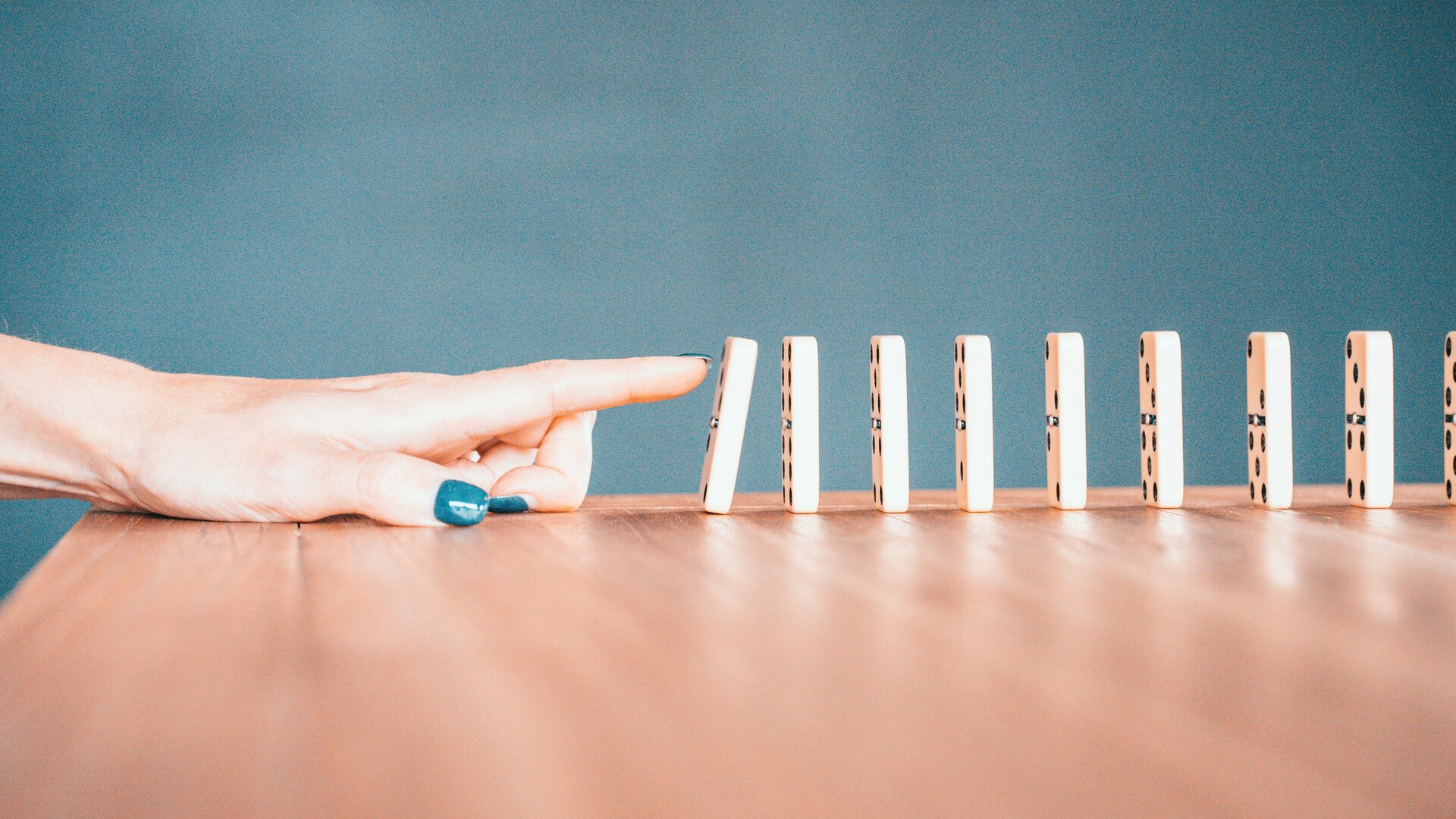 Auf dem Bild ist eine Hand mit blau lackierten Fingernägeln zu sehen, ein Finger ist dabei Dominosteine umzustoßen | © Bradyn Trollip/Unsplash